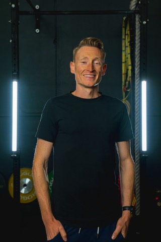 Smiling man in black T-shirt standing in a gym with fitness equipment and vertical lights behind him.