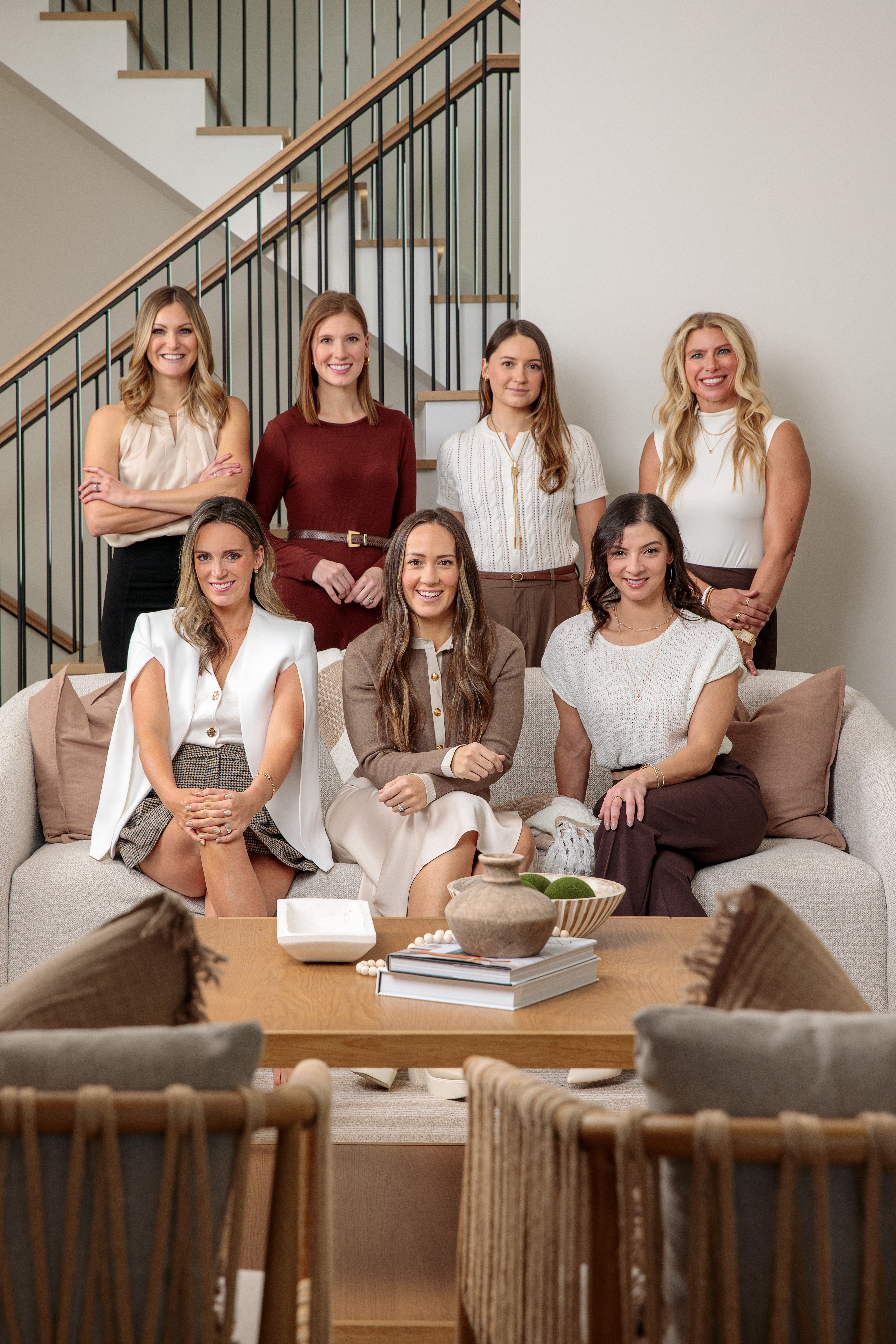Seven women pose in a living room, some sitting on a sofa and others standing behind, smiling at the camera.