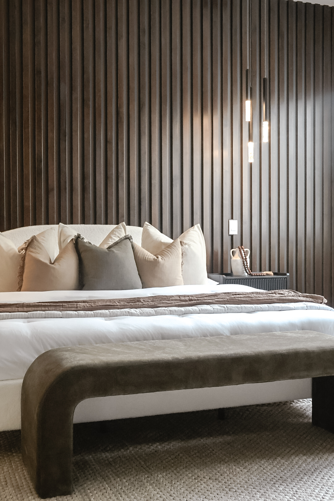 Bedroom with dark wooden wall, bed with beige and gray pillows, and a bedside lamp.