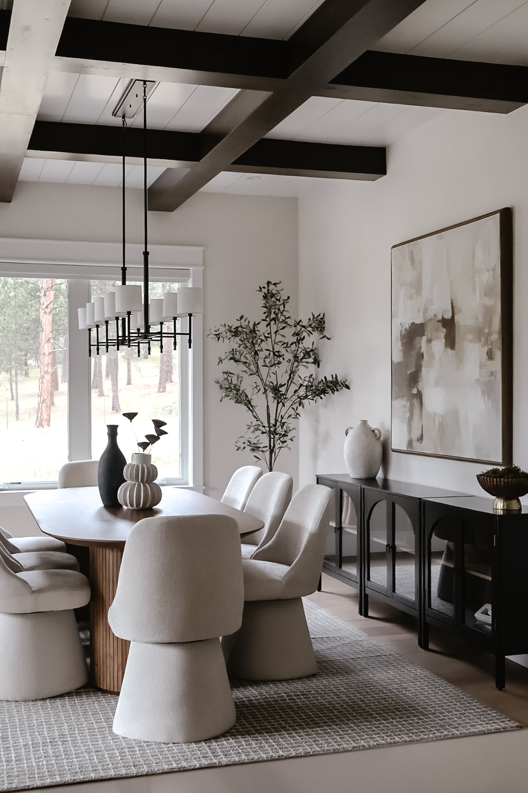 Dining room with white chairs around a wooden table, black ceiling beams, large window, and abstract wall art.