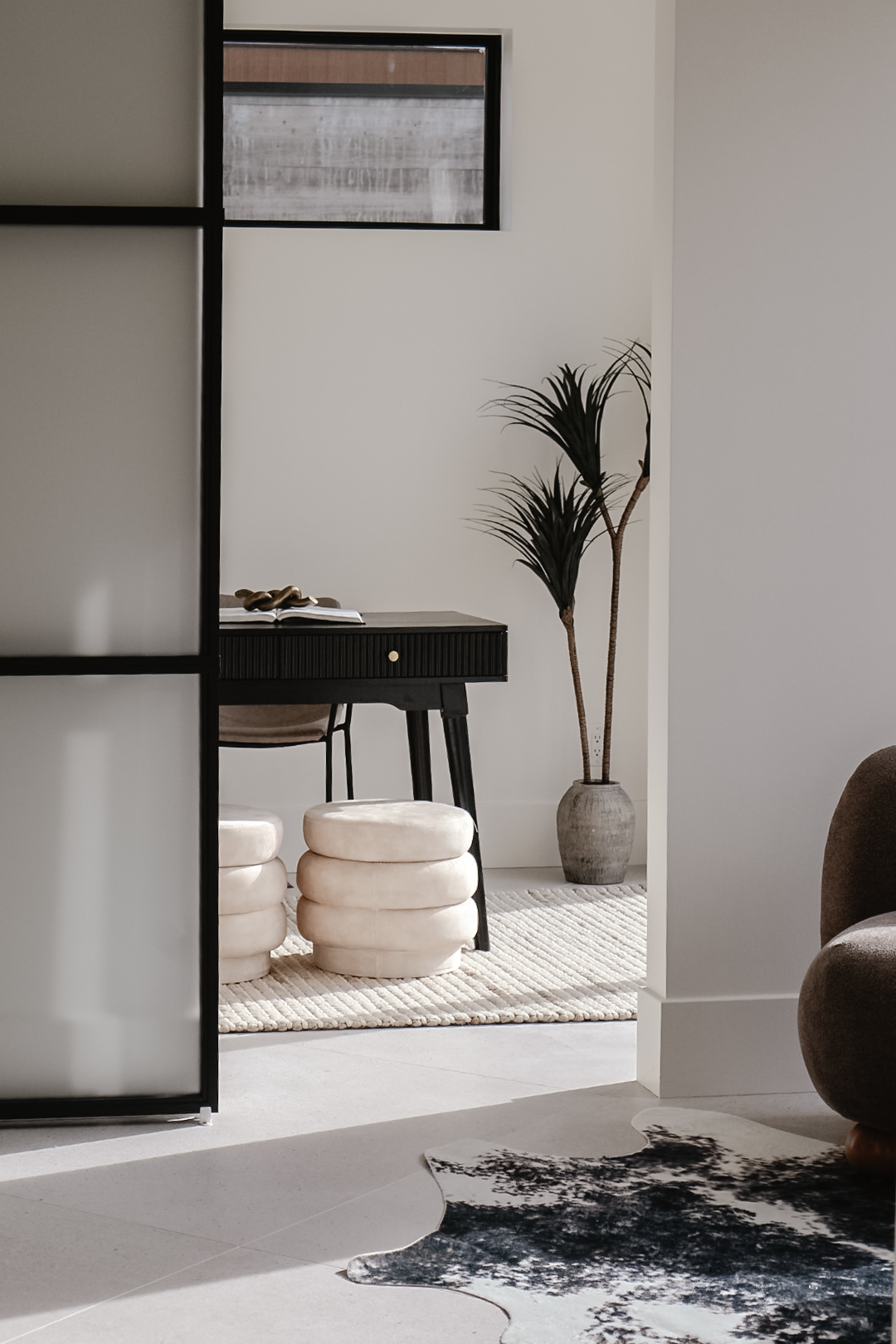 Interior room with a black desk, potted plant, and stacked white cushions near a white wall and window.
