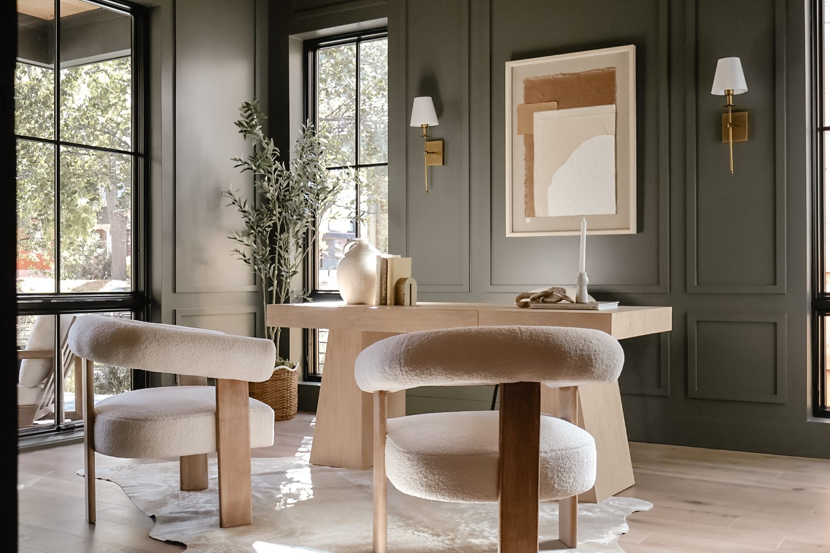 Dining room with two upholstered chairs, a round table, and wall art, with large windows and green walls.