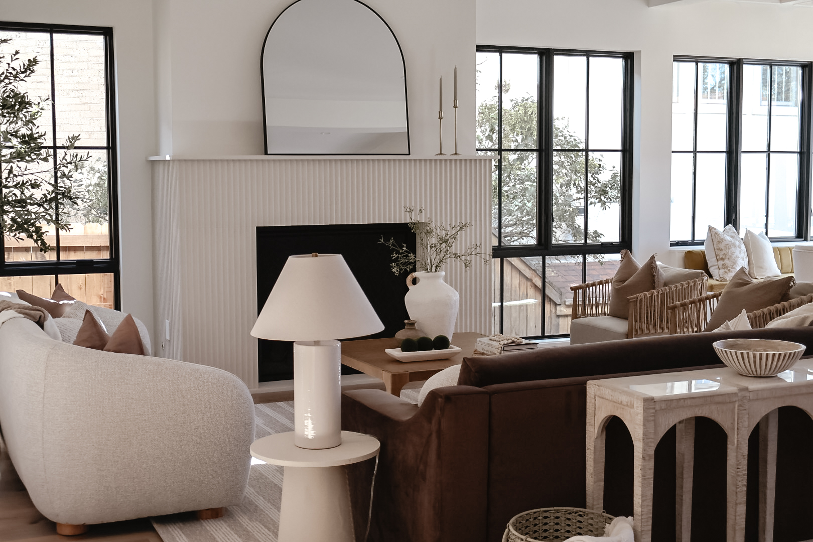 Living room with large windows, white fireplace, mirror, sofa, armchair, table lamp, and decorative items.