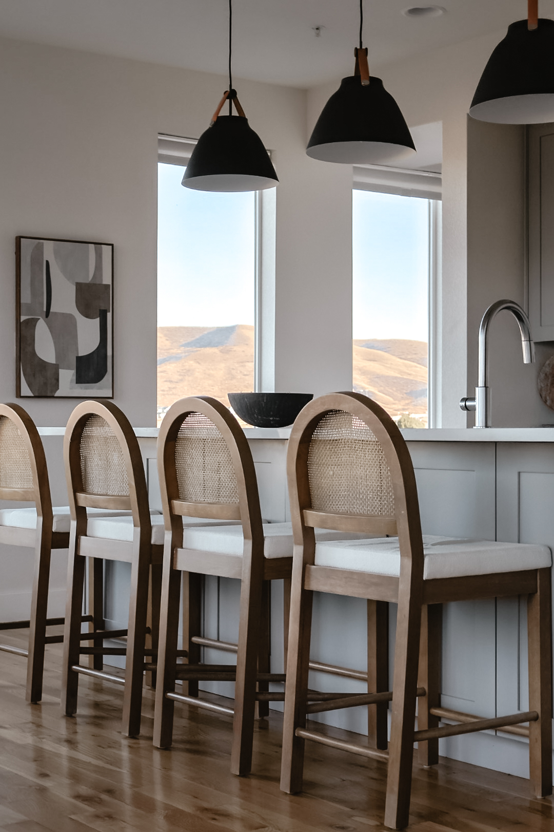 Kitchen with four bar stools at a counter, large windows showing landscape, pendant lights hanging, and a bowl on the counter.