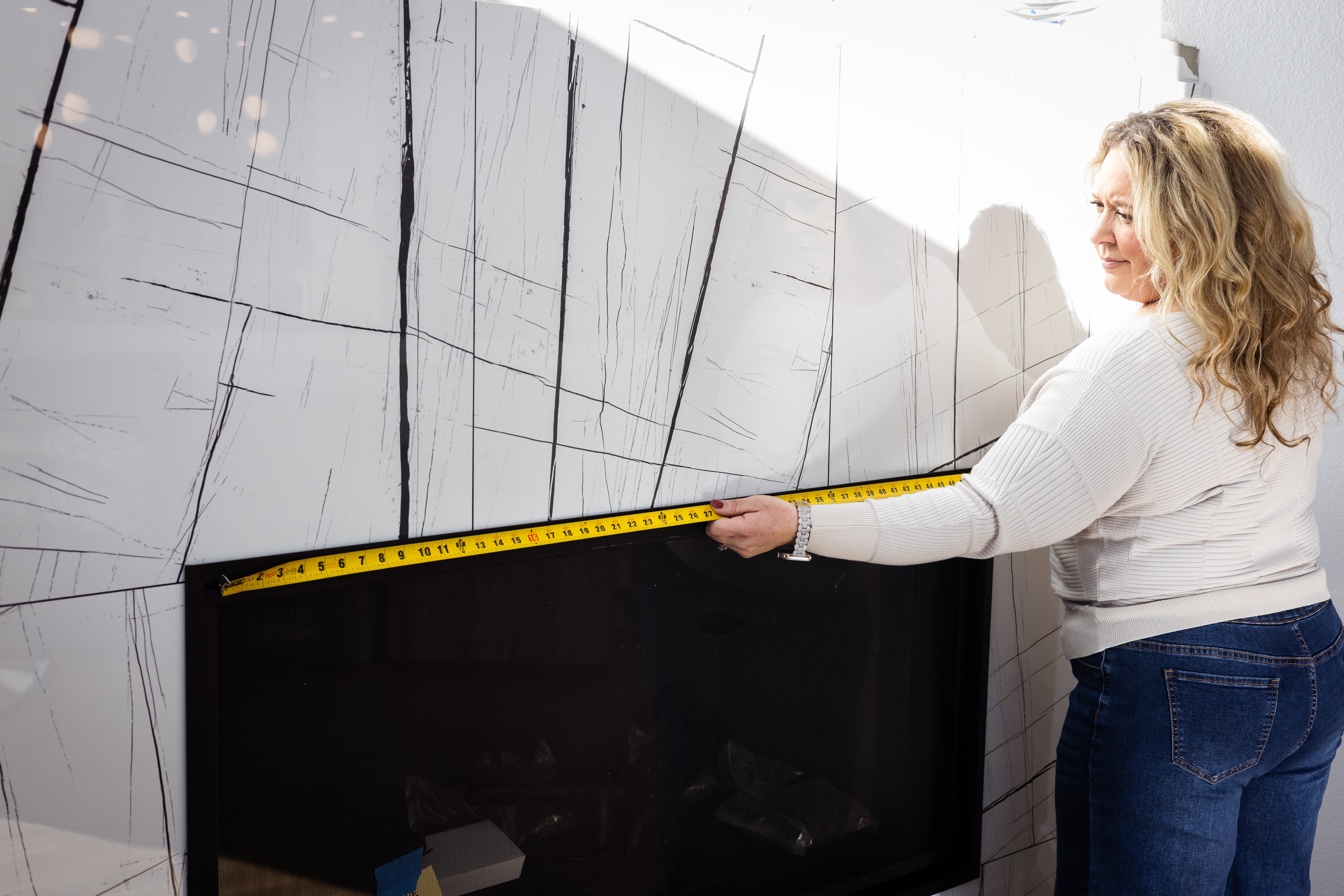 Woman measuring a large marble wall with a yellow tape measure indoors.