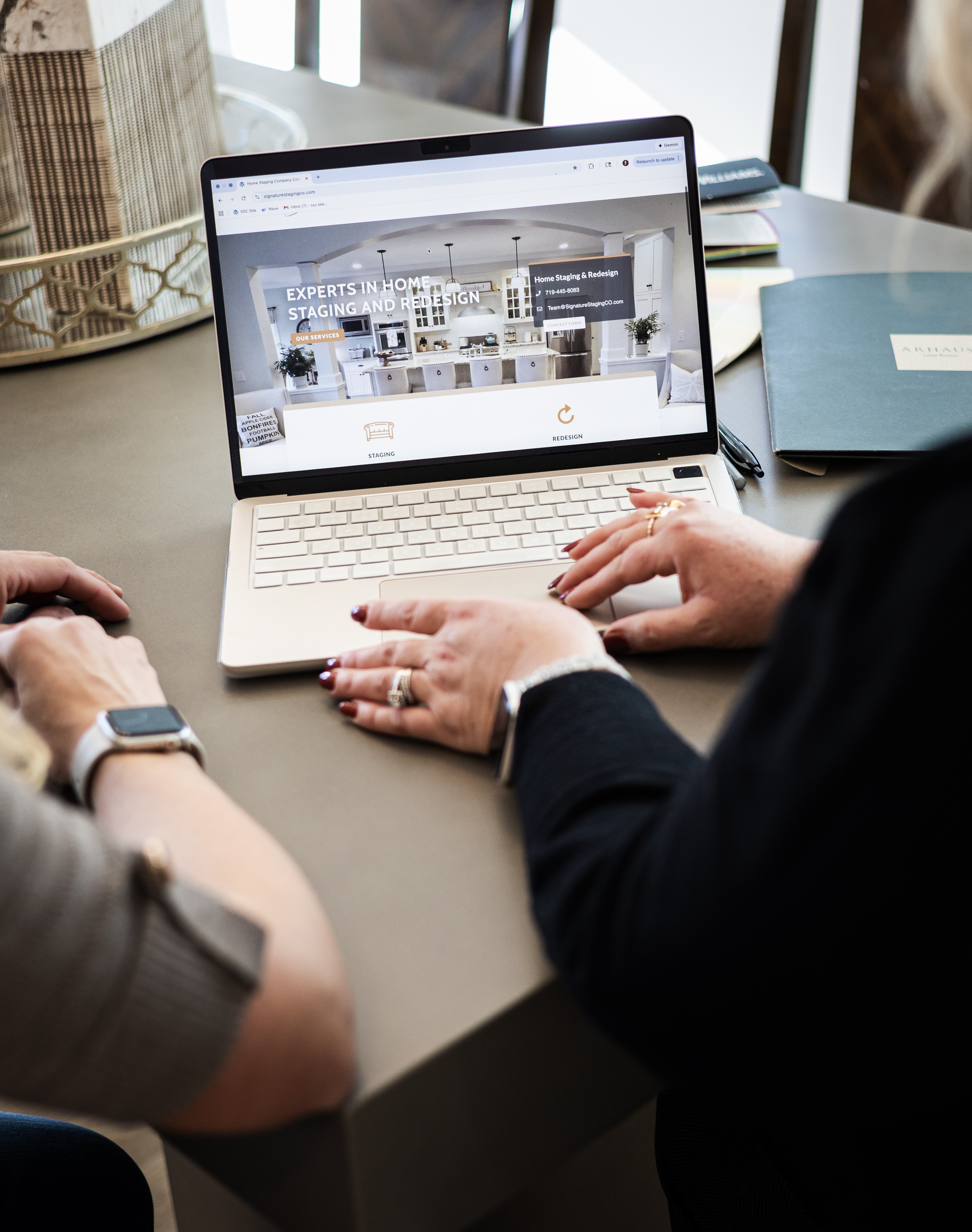 Two people discussing a laptop displaying a website, with notebooks and a plant on the table.
