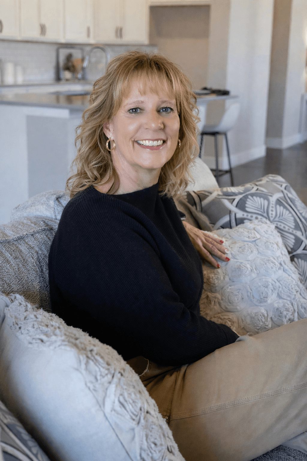 Woman with blonde hair smiling, sitting on a couch in a living room, wearing a black top.