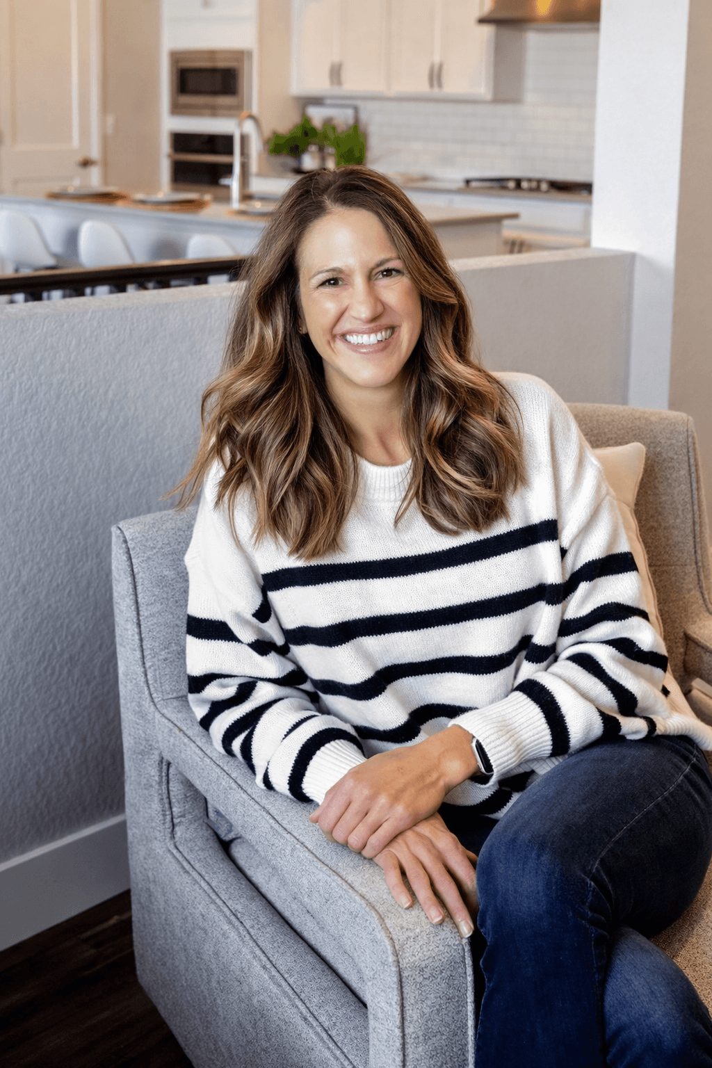 Woman with long wavy hair smiling, sitting on a gray armchair in a modern kitchen.
