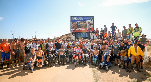 Large group of people, including children and adults, gathered outdoors with some in wheelchairs, under a blue sky.