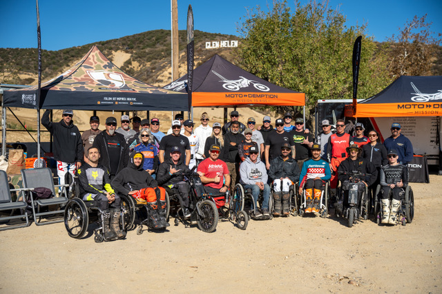 Group of people with motorcycles and wheelchairs outdoors, tents in background, sunny day, hilly landscape.