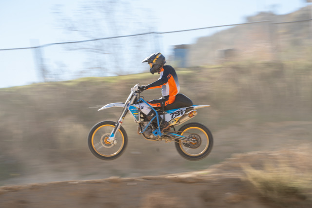 Person riding a dirt bike in mid-air, wearing a helmet and orange and black gear, on a dirt track.