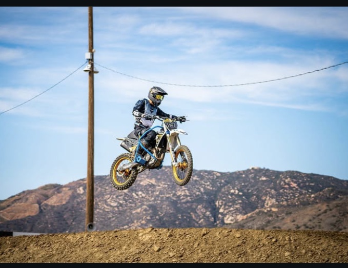 Motocross rider in gear jumps over dirt track with mountains and blue sky background.