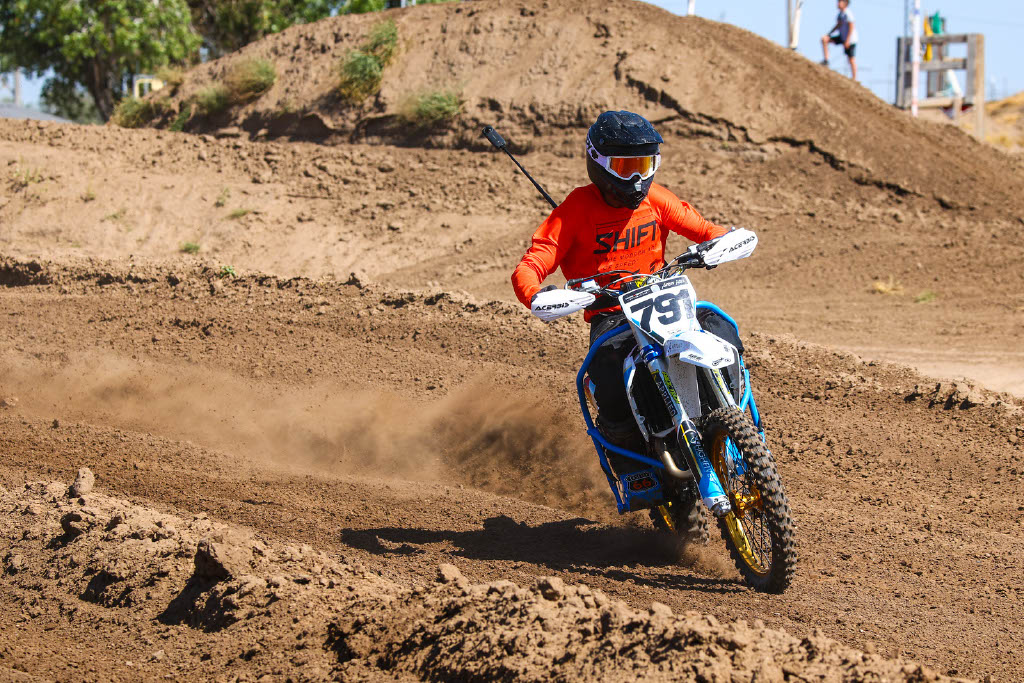 Person riding a dirt bike on a dirt track, wearing a helmet and orange shirt, kicking up dust.