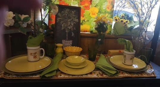 Table setting with yellow plates, cups, green napkins, and flower arrangements in the background.