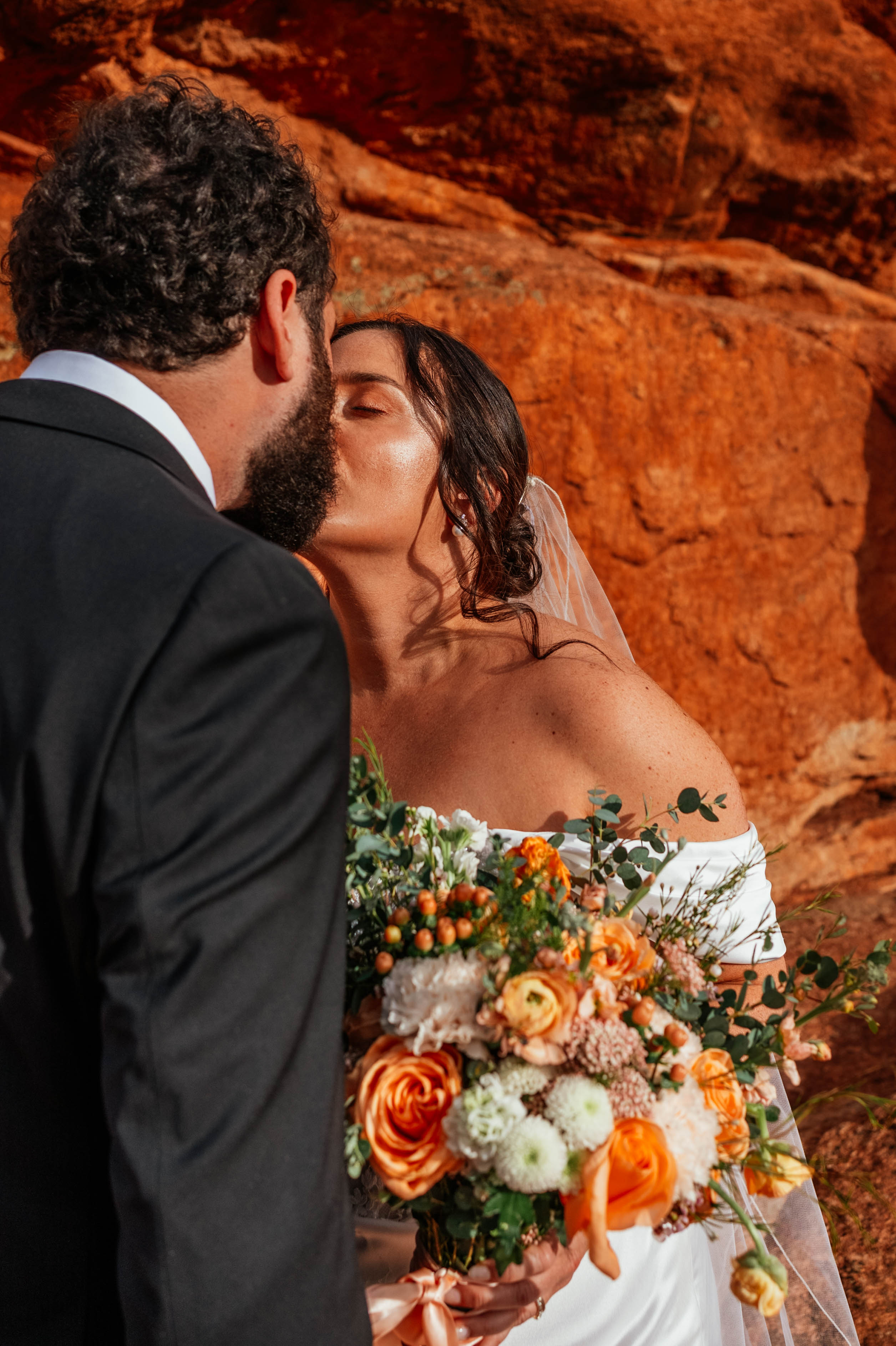 Bride and groom share a kiss outdoors with a large bouquet of flowers, red rocks in the background.