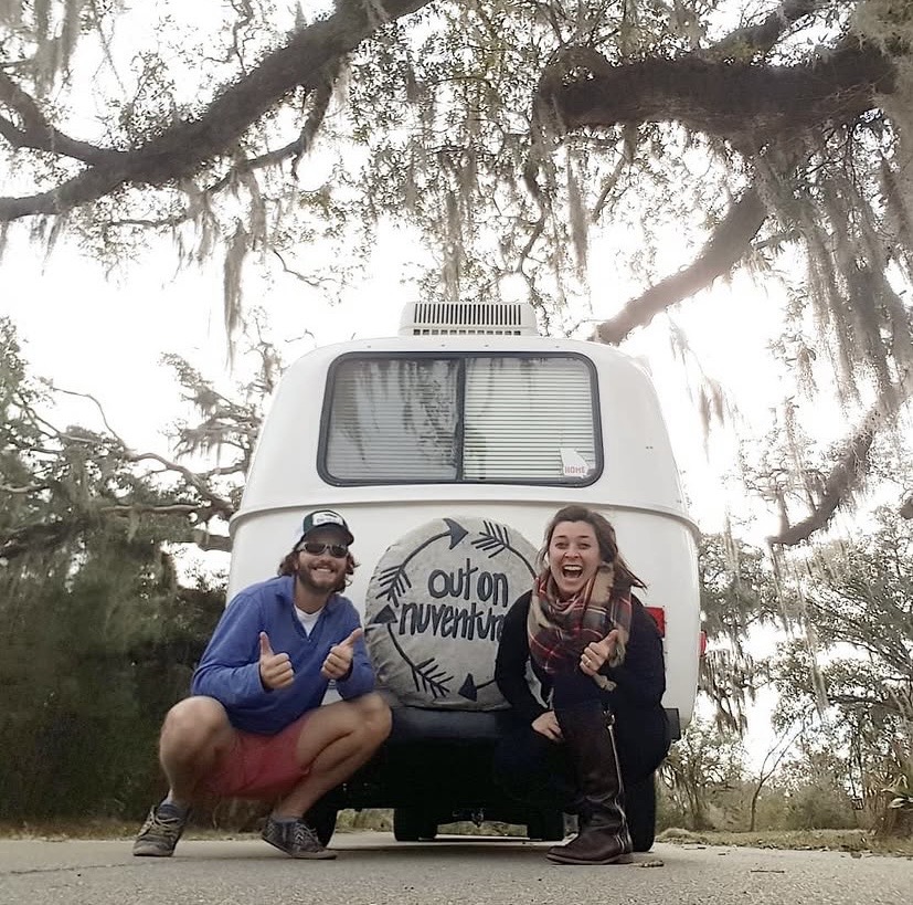 Two people crouch in front of a camper van, smiling and giving thumbs up, with trees and water in background.