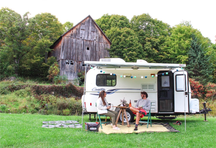 Two people sitting outside a white camper trailer on a grassy field with a barn and trees in background.