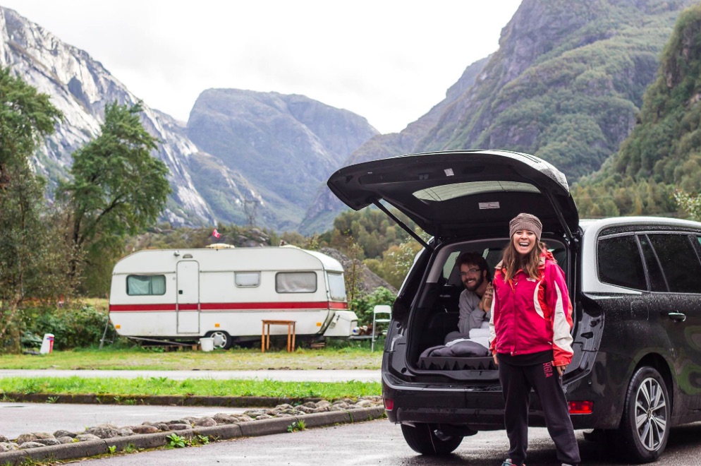 Woman standing next to an open car trunk with a mountain landscape in the background.