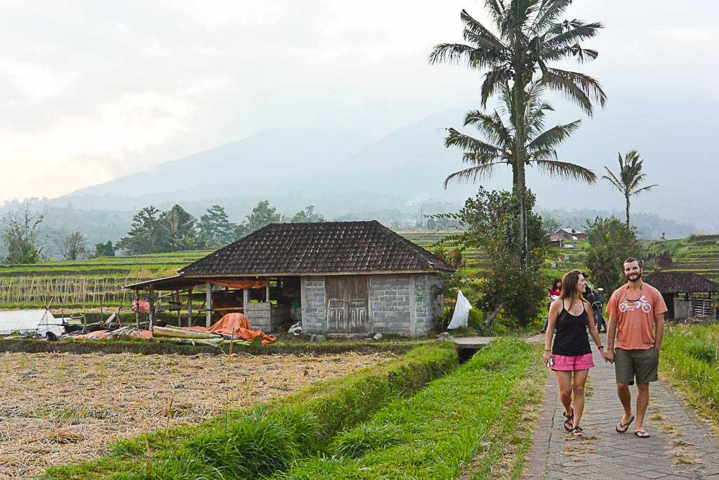 Two people walk along a path in a rural area with a house, palm trees, and mountains in the background.