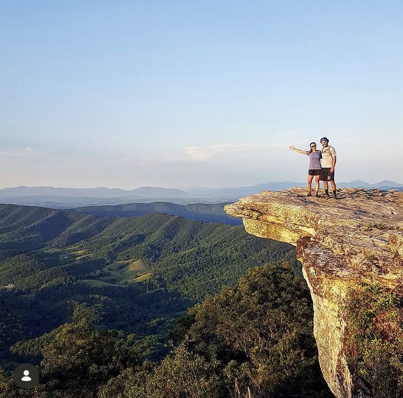Two people stand on a rocky cliff edge overlooking a green valley and distant mountains under a clear blue sky.