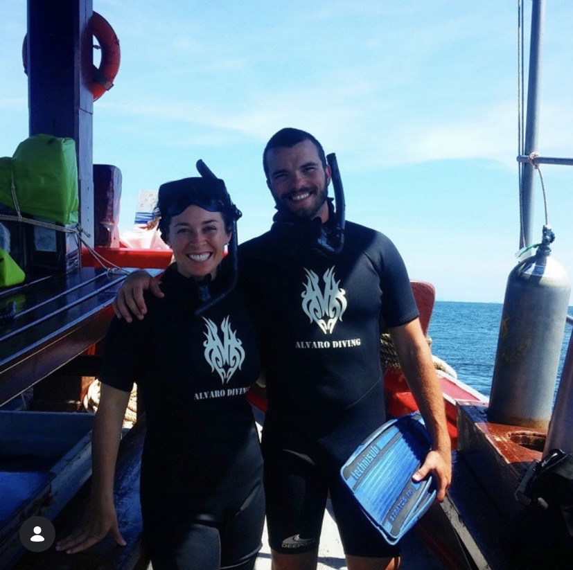 Two smiling people in wetsuits on a boat, with the ocean and sky in the background.