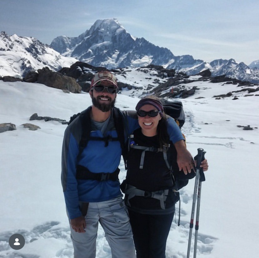 Two hikers in snow-covered mountains, smiling, wearing backpacks and sunglasses, with a mountain peak in the background.