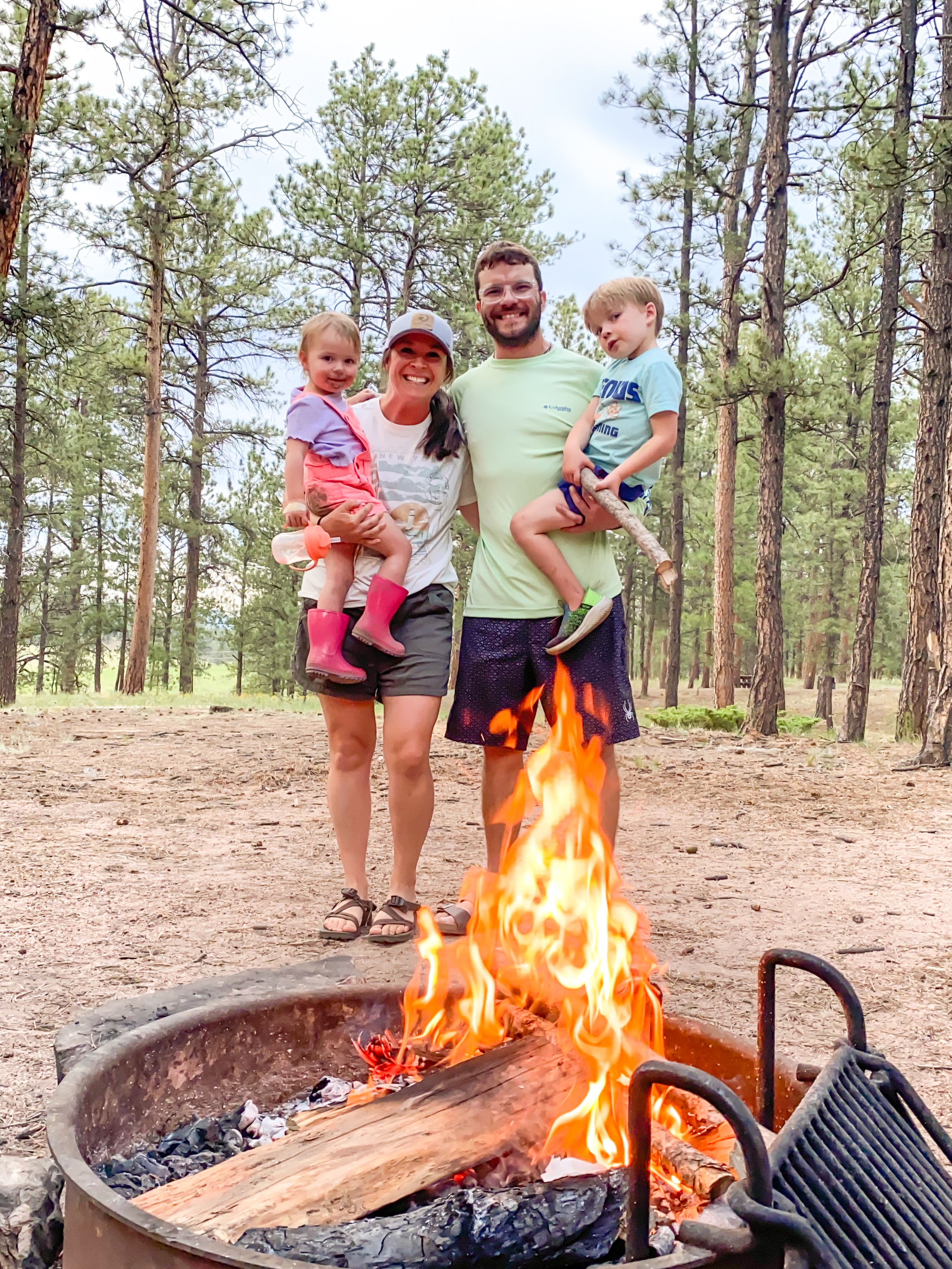 Group of four people, two children and two adults, standing outdoors near a campfire in a forest.