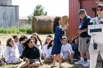 Group of children sitting outdoors, girl standing and talking to a woman with a clapperboard, hay bale in background.