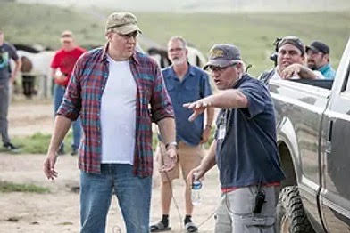Two men in conversation outdoors with several people and vehicles in background.