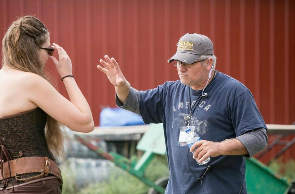 Man wearing glasses, cap, and navy shirt gestures with his hand while talking to a woman with long hair and sunglasses.