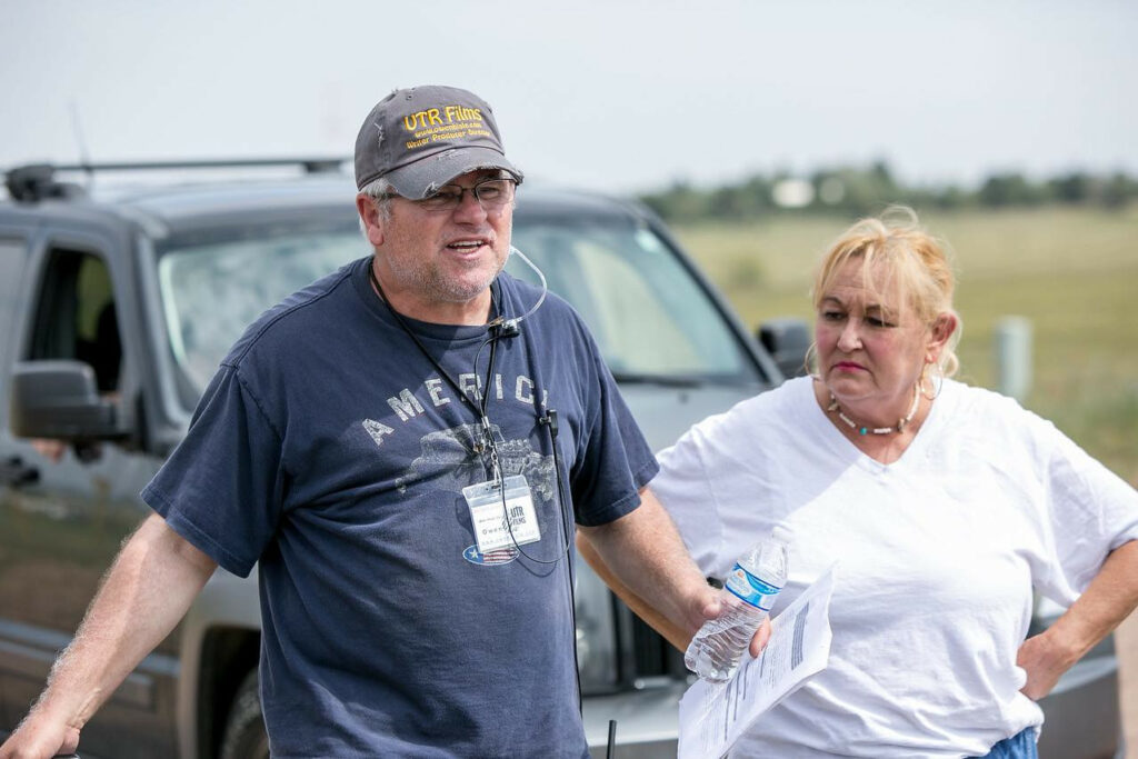 Two people standing outdoors near a vehicle, one holding a water bottle, the other with hands on hips, in a field.