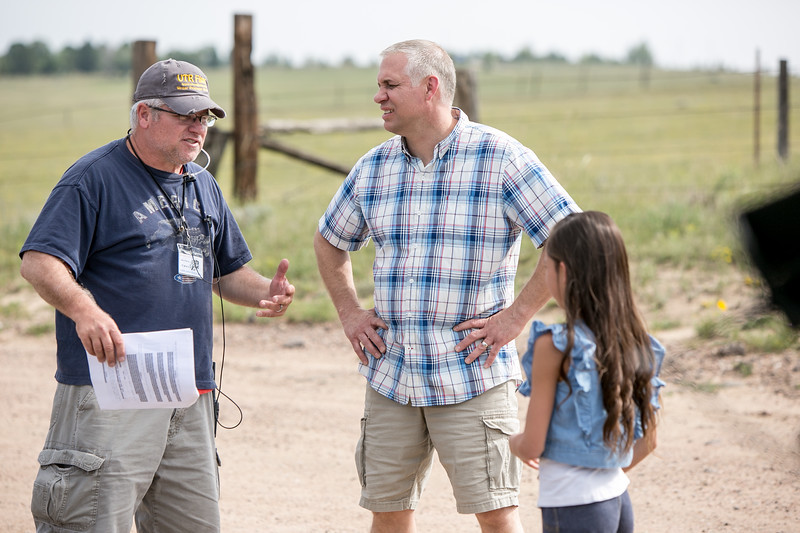 Three people, two men and a girl, standing outdoors on a dirt path with a fence and field in background, talking.