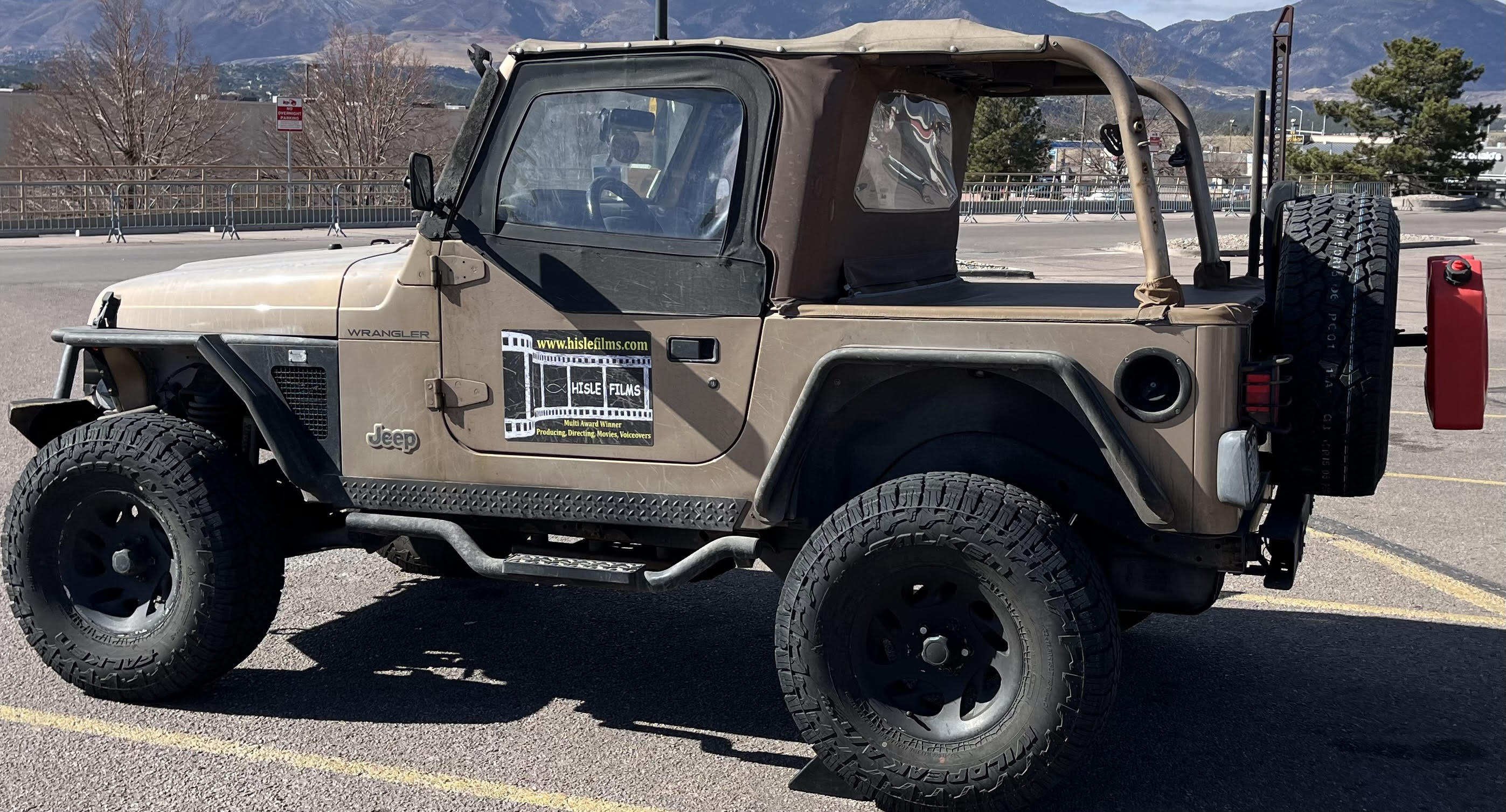 Beige off-road vehicle with a spare tire on the back and a red object attached to the rear, parked on a road.