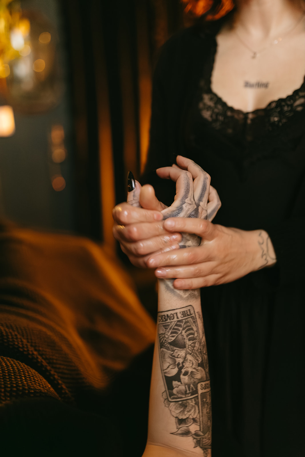 Person with tattooed arm holding another person's hand, wearing a black lace dress, in a dimly lit setting.
