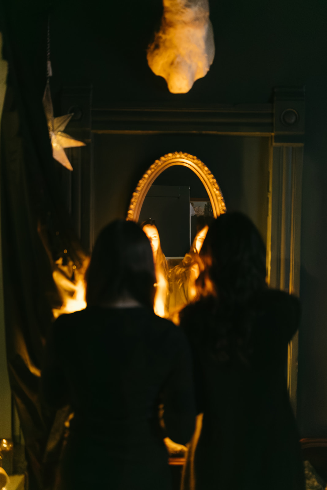 Two people with dark hair and dark clothing face a mirror with a golden frame, illuminated by warm light in a dim room.