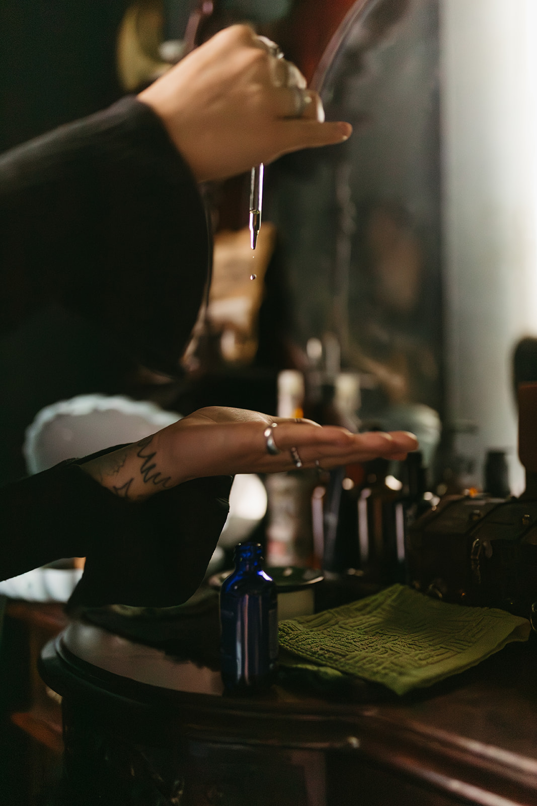Person's hand with rings and tattoo, reaching towards a mirror with various bottles and objects on a table.