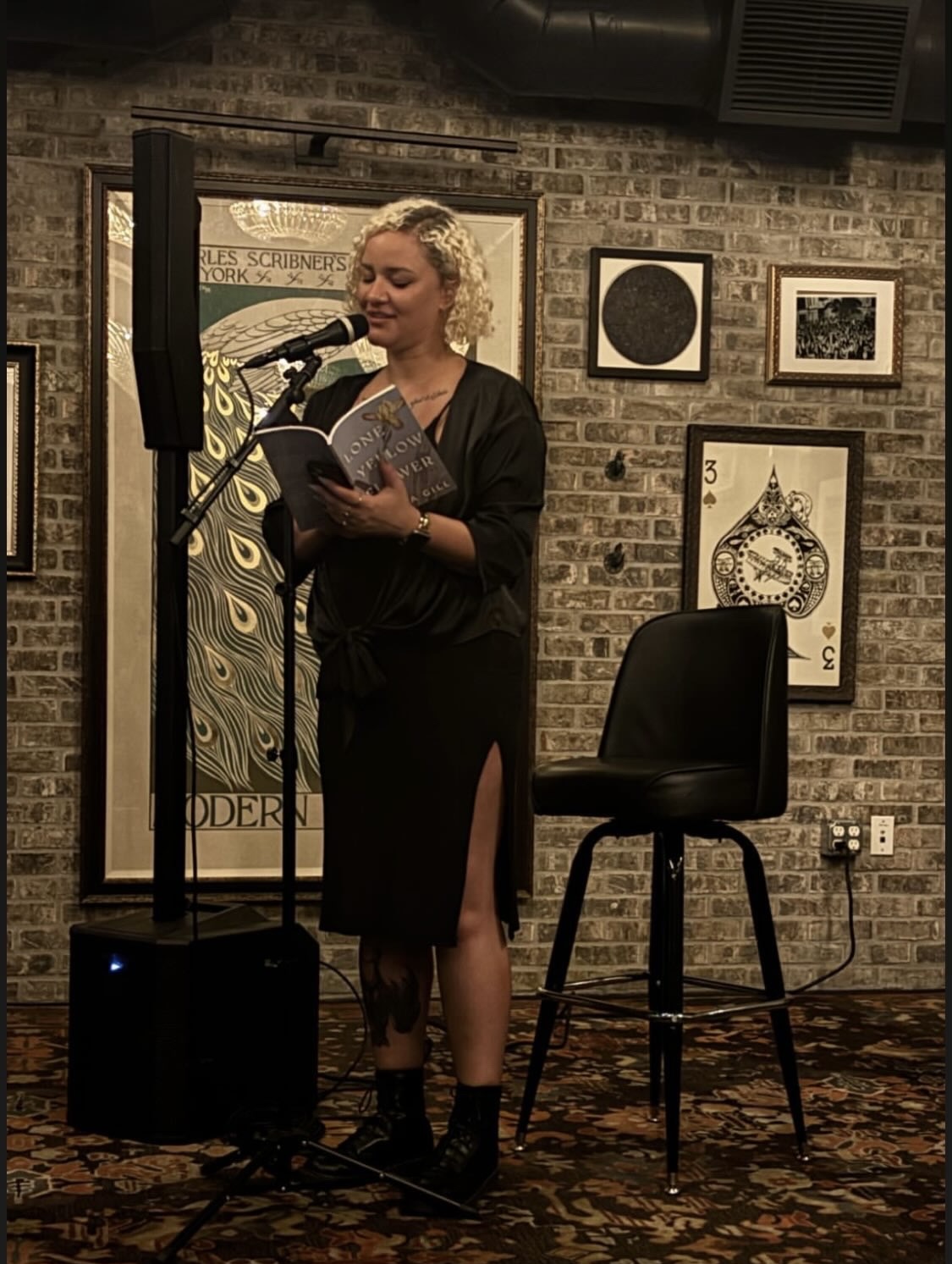 Woman with curly blonde hair reads from a book into a microphone on stage, brick wall background with framed artwork.