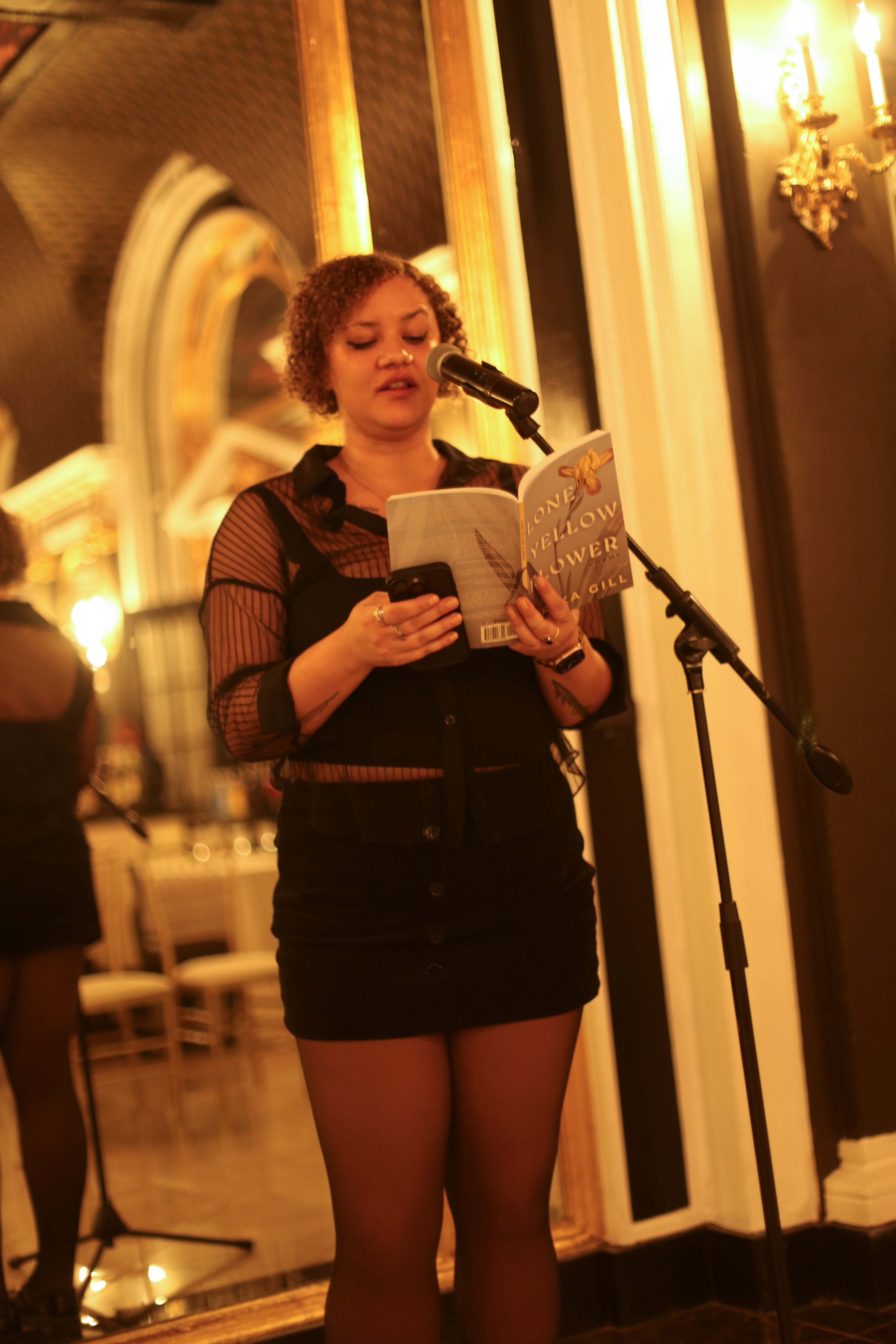 Young woman reading from a book at a microphone in an elegant room with ornate decor and warm lighting.