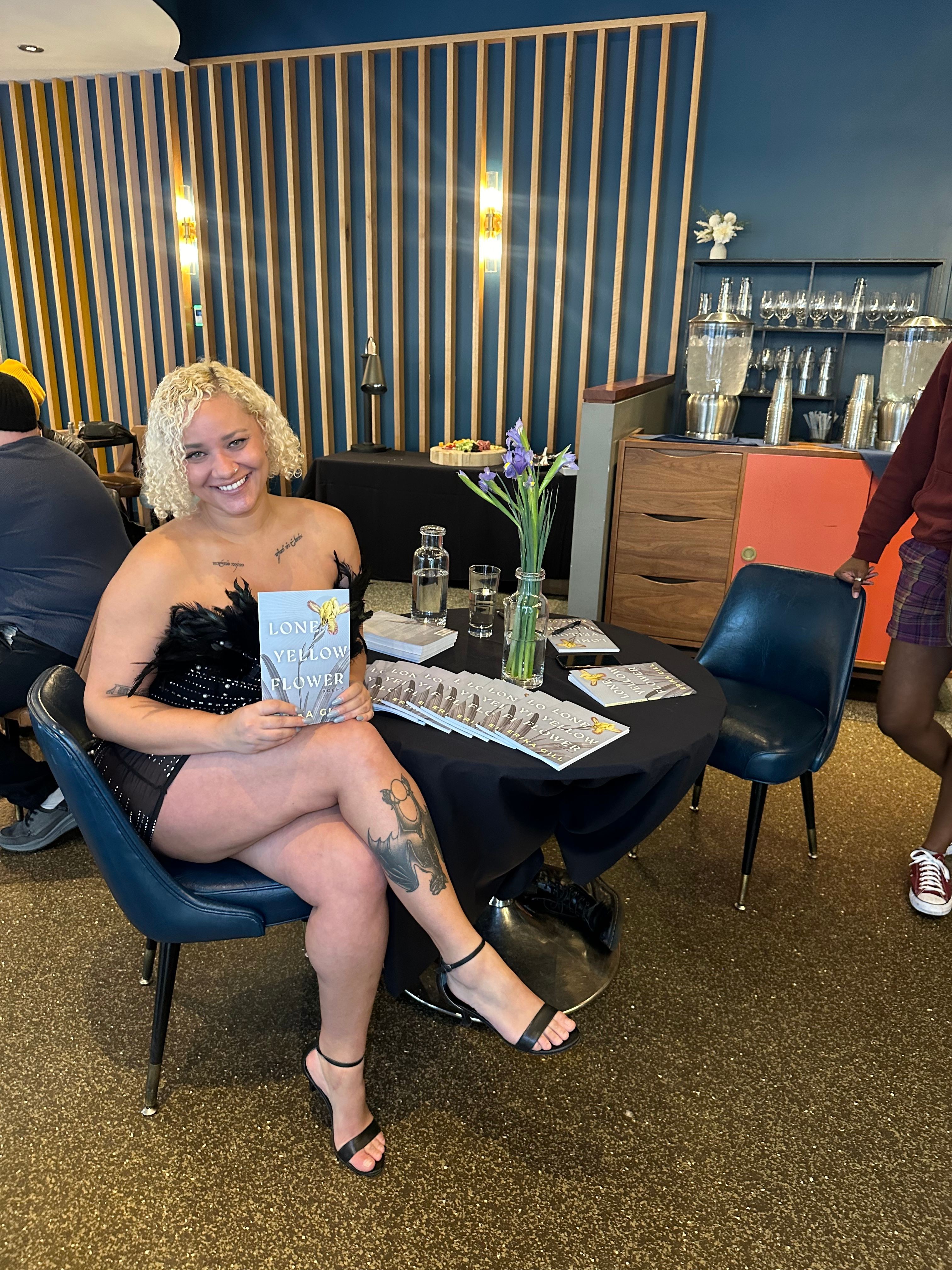 Woman with blonde hair sitting at a table, smiling, with a tattoo on her leg, in a decorated indoor setting.
