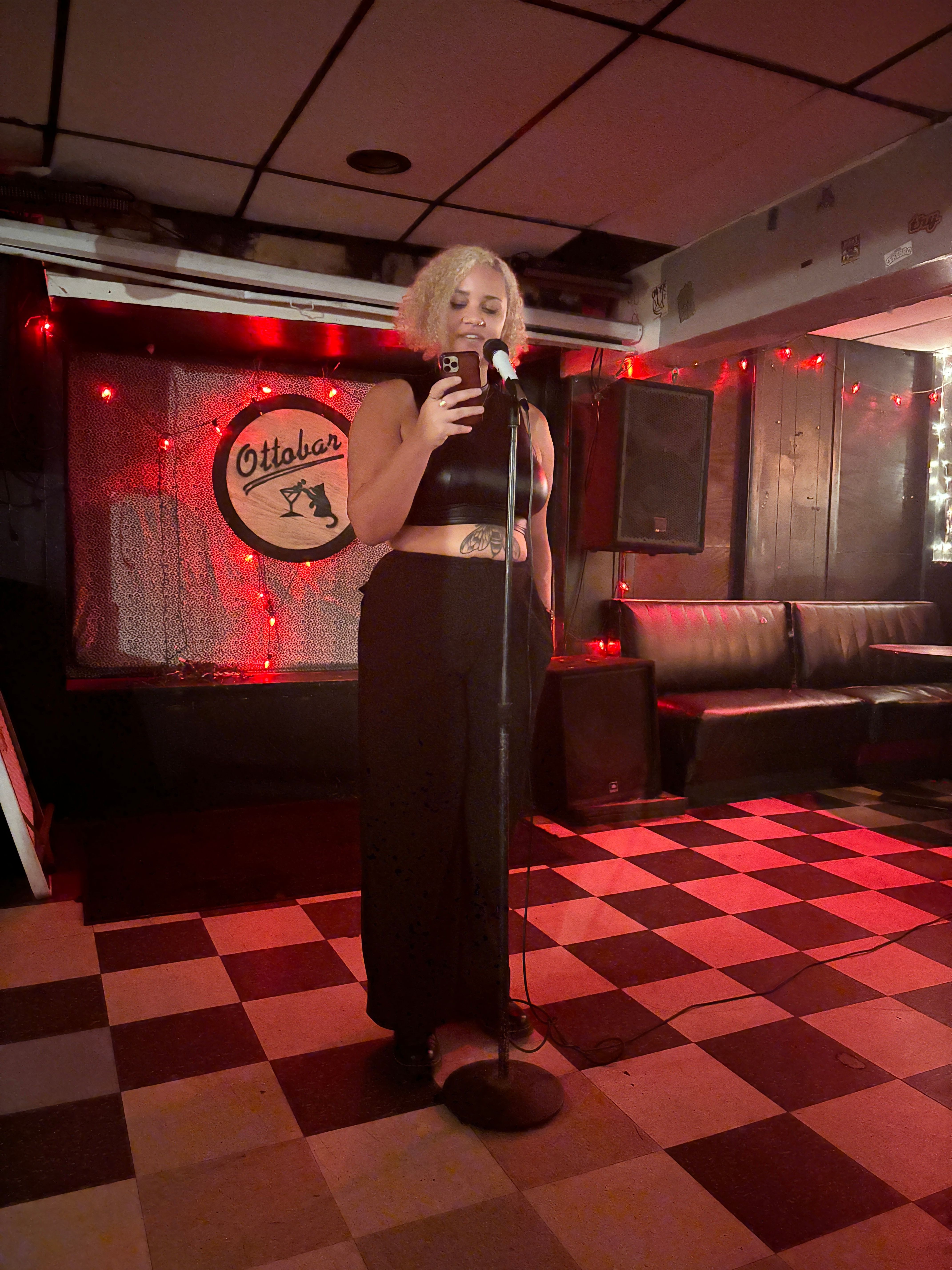 Woman with blonde hair singing into a microphone on stage with red lighting and a checkered floor.