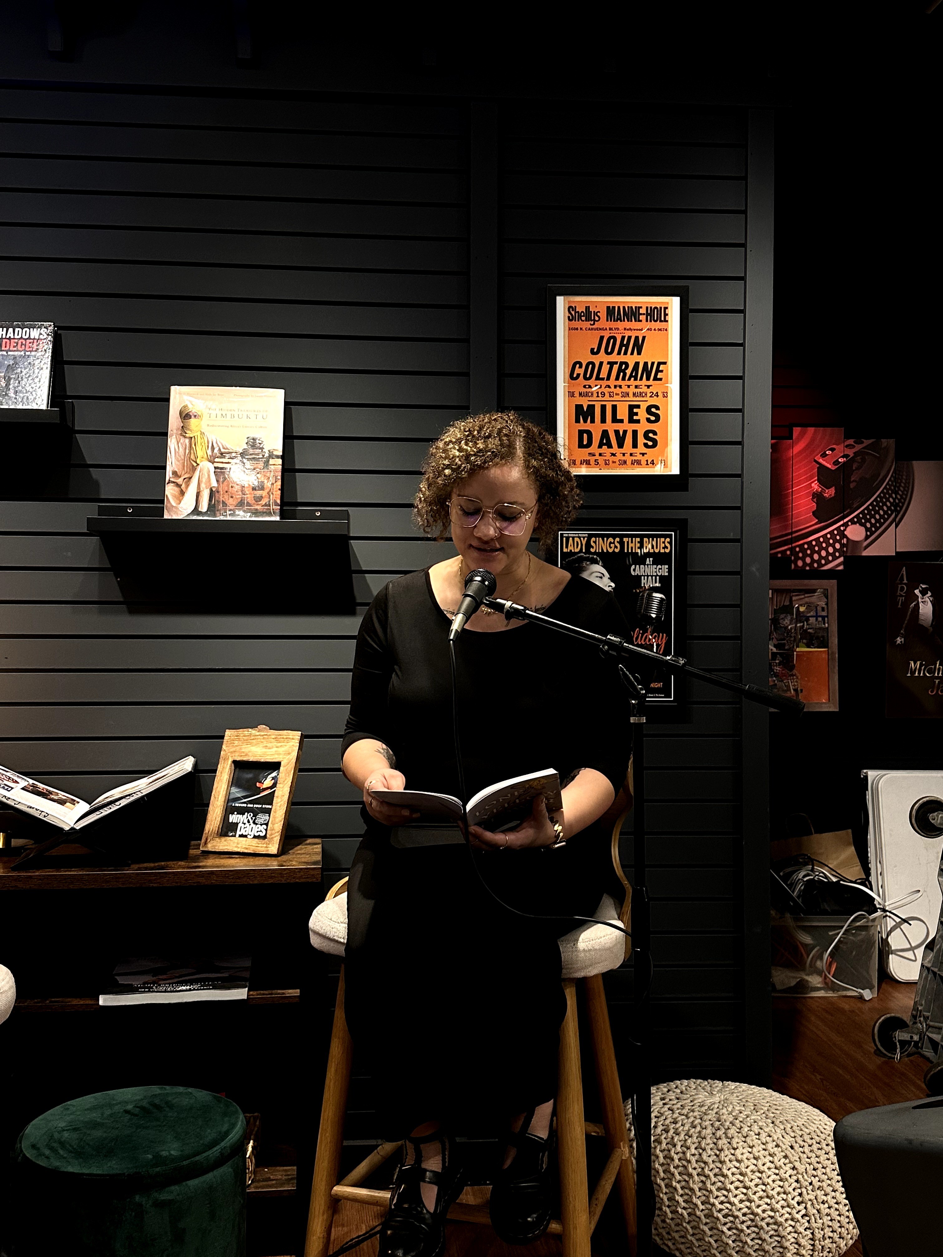 Woman with curly hair reading a book and speaking into a microphone in a room with black wall decor.