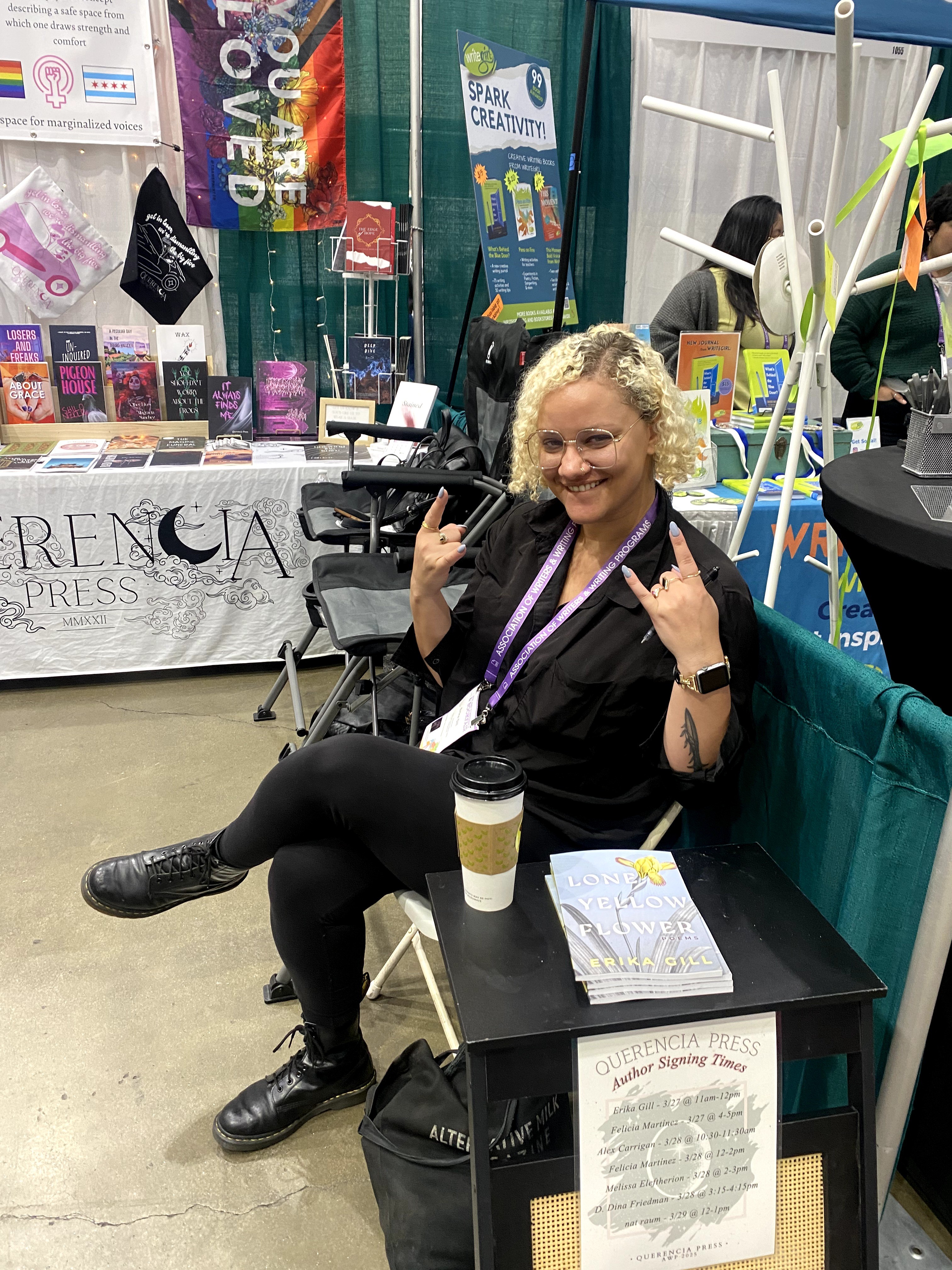 Woman with curly blonde hair sitting at a table, smiling, holding a pen, with books and banners in the background.