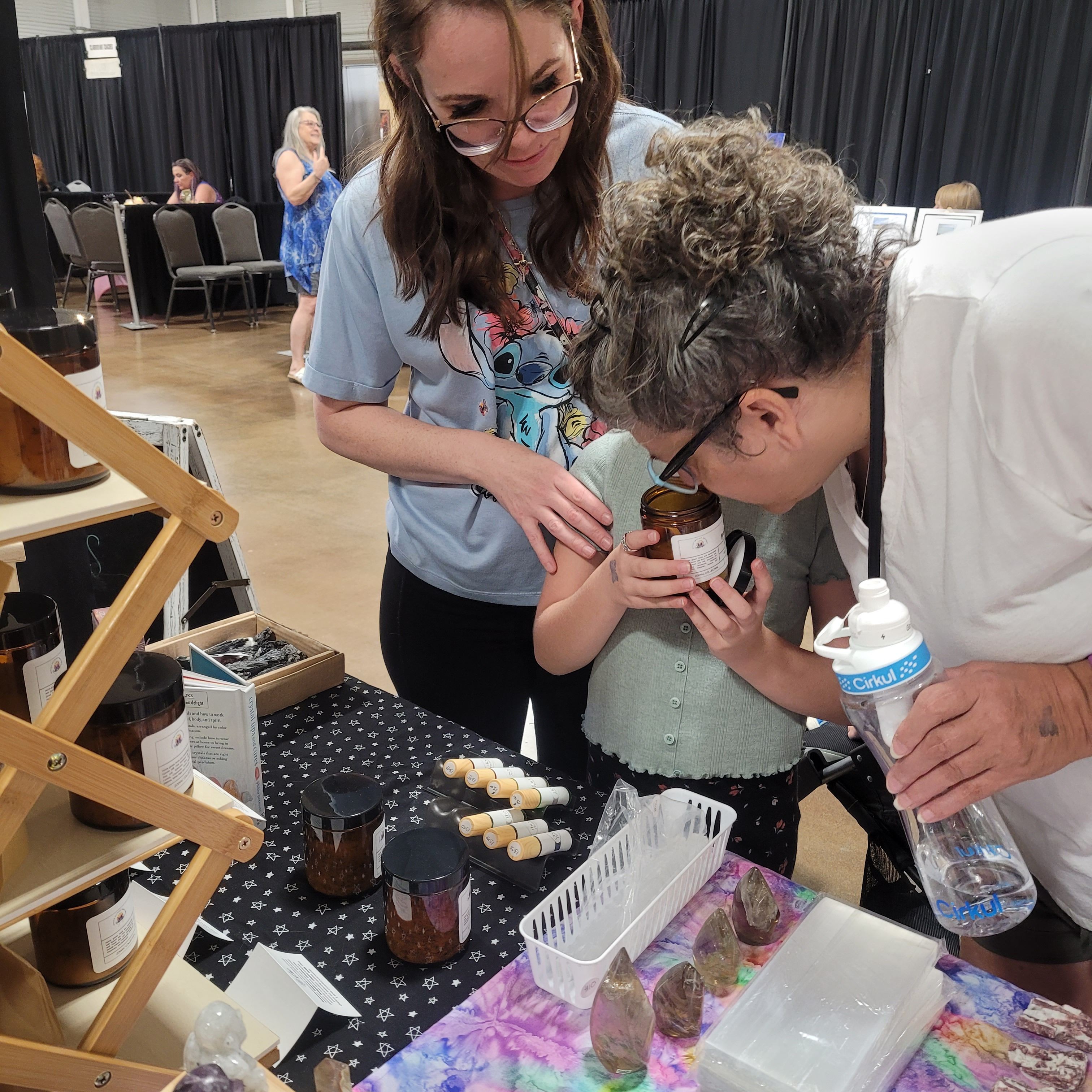 Two women and a child examining a small object at a table with various items, in an indoor event space.