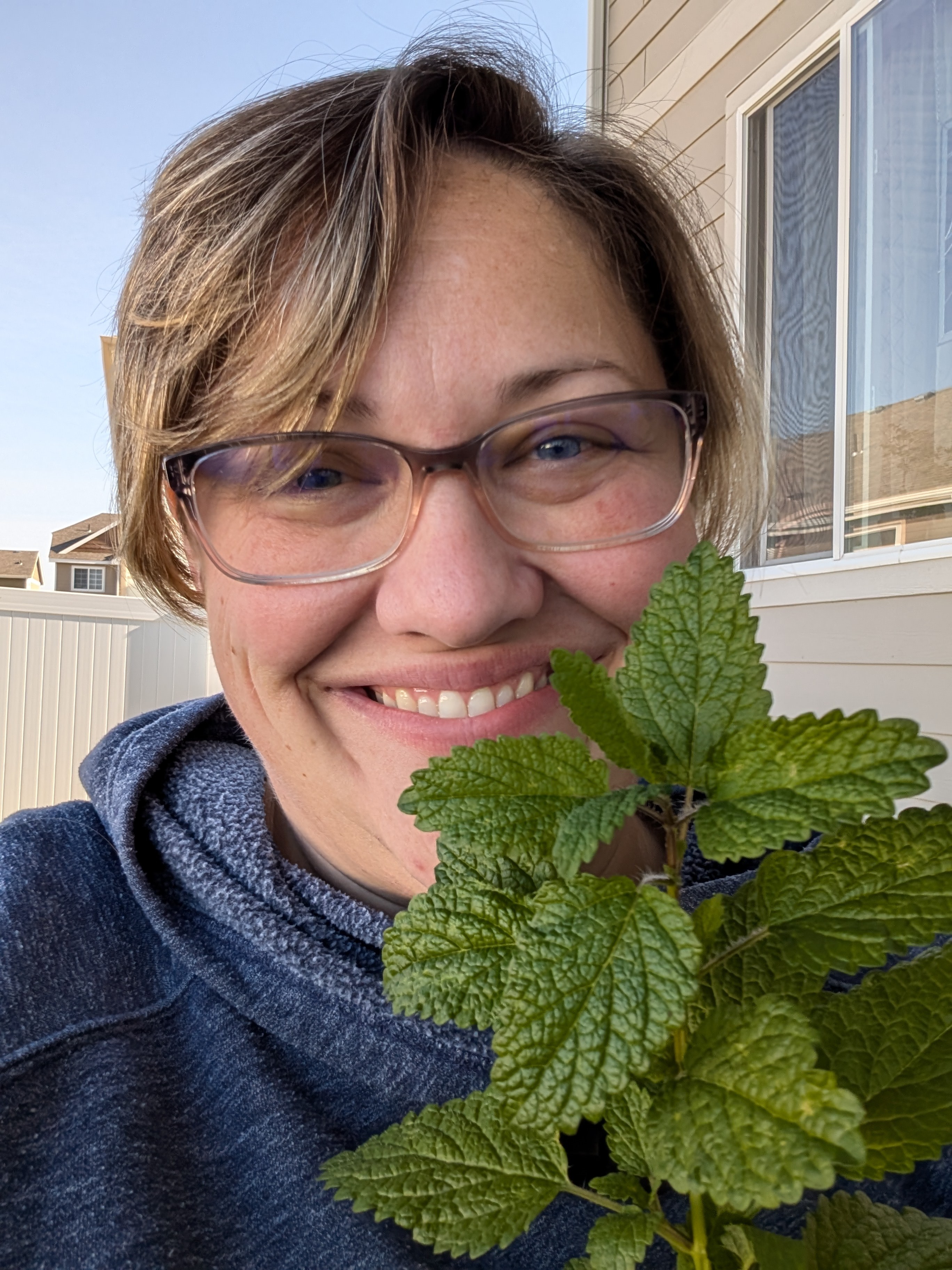 Woman with glasses smiling outdoors near a house, holding a green leafy plant.