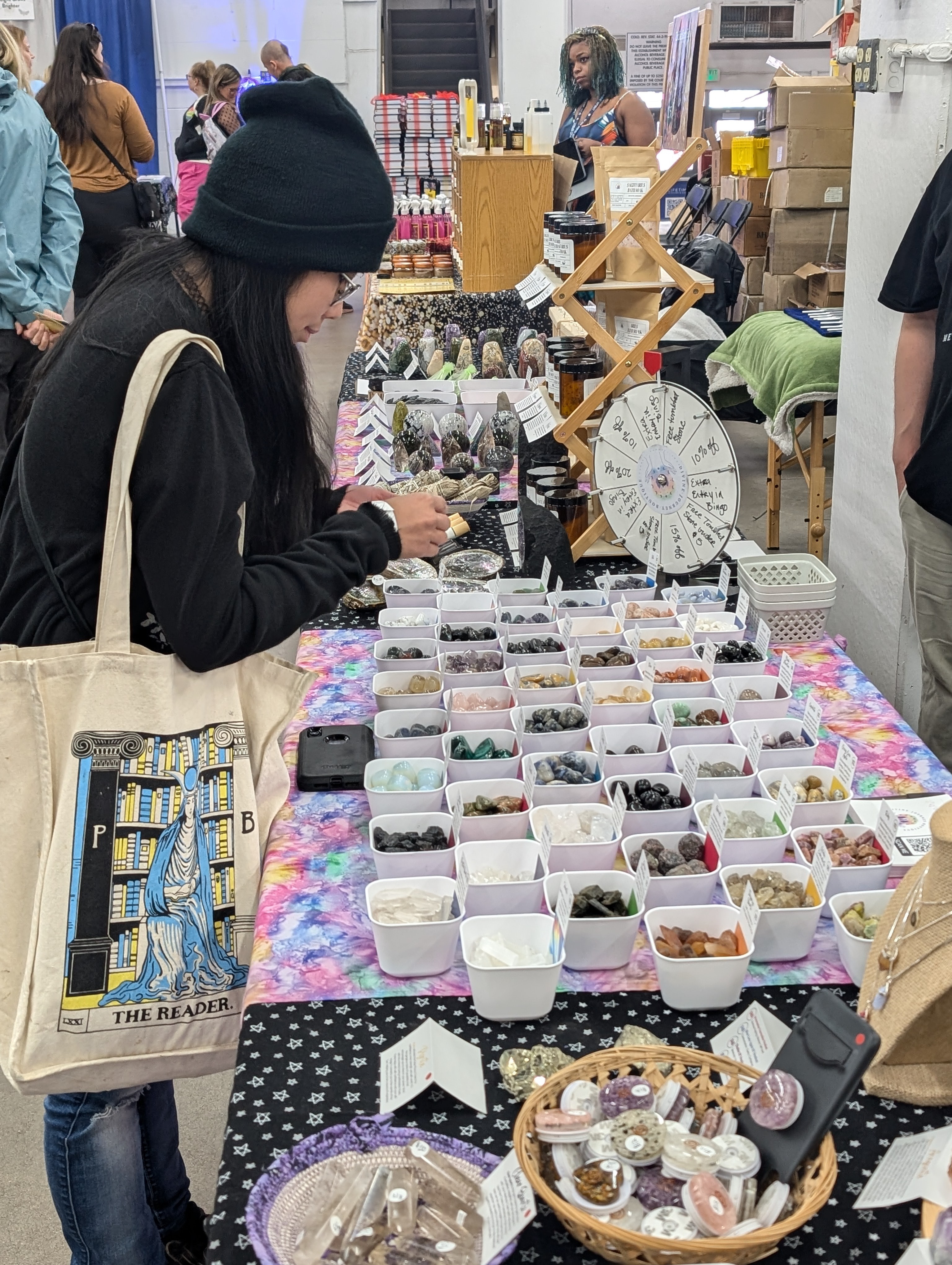 Person browsing jewelry at a market stall with various accessories in containers, other shoppers in background.