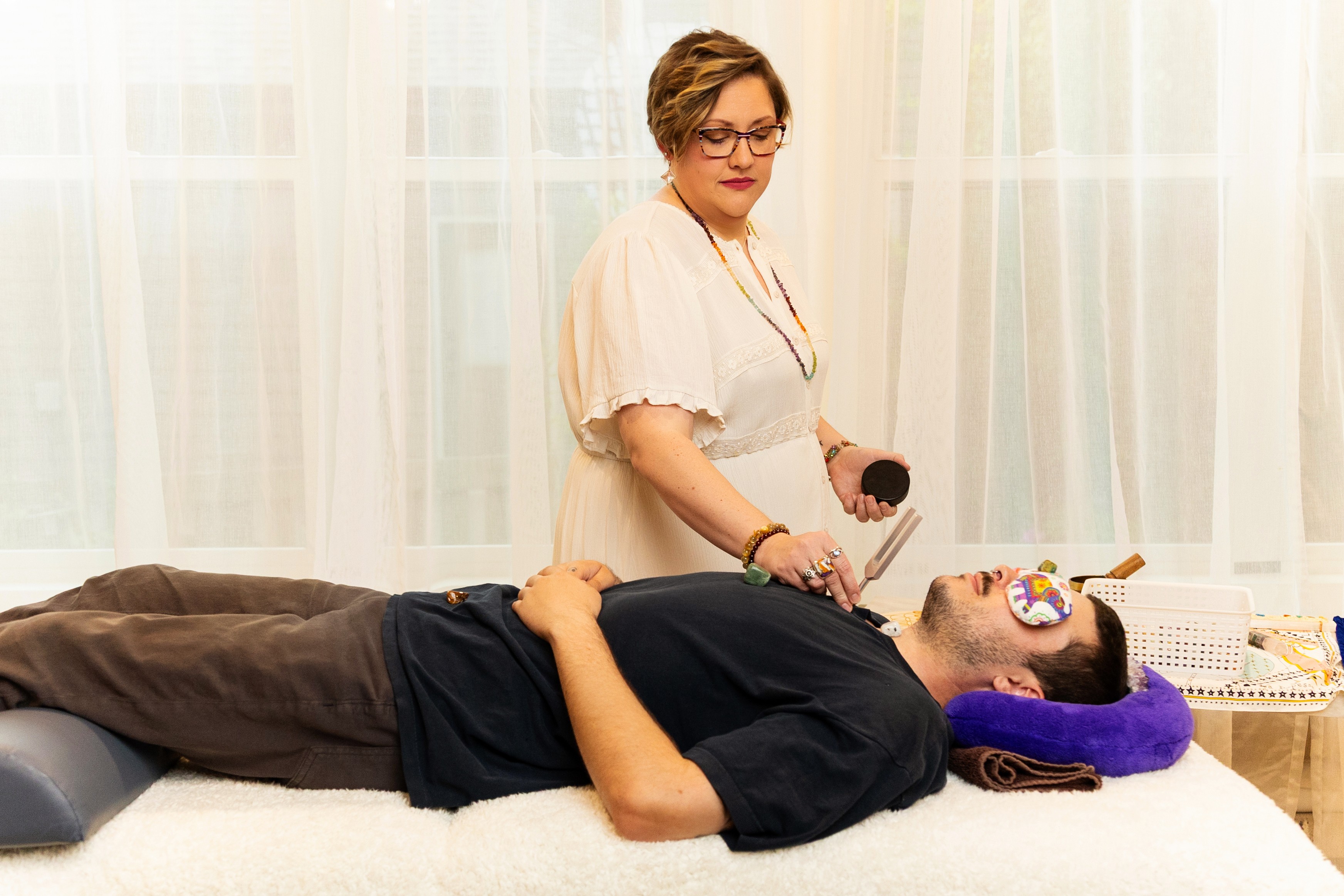 Woman with glasses and short hair stands beside a man lying on a bed, holding a tool, in a bright room.