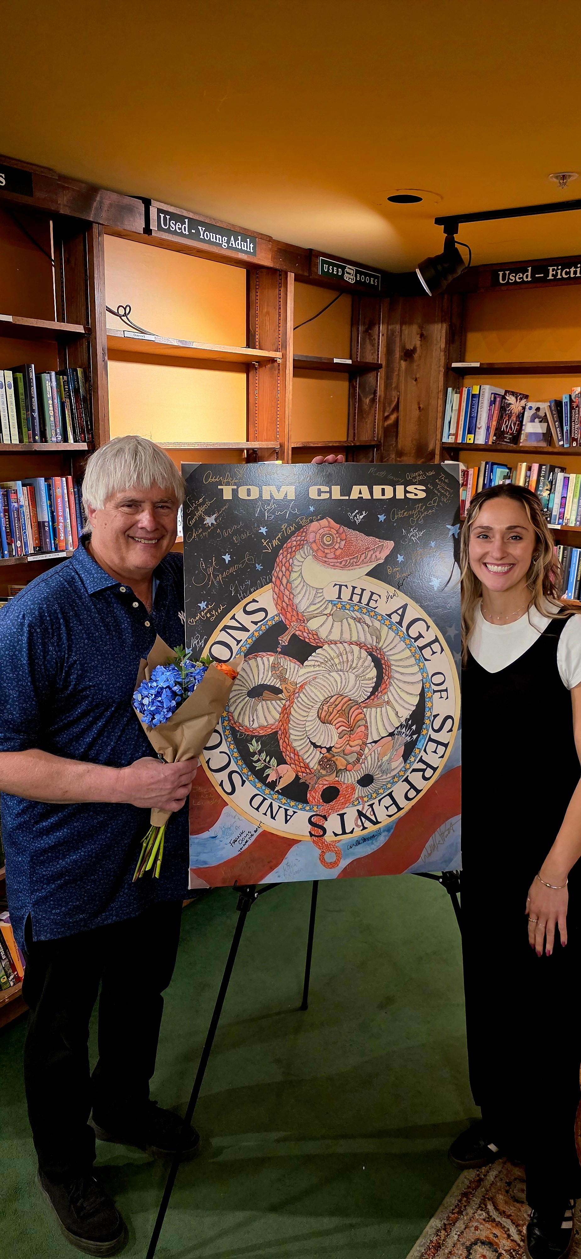 Two people standing in a library, holding a large poster with a snake illustration and text, bookshelves in the background.