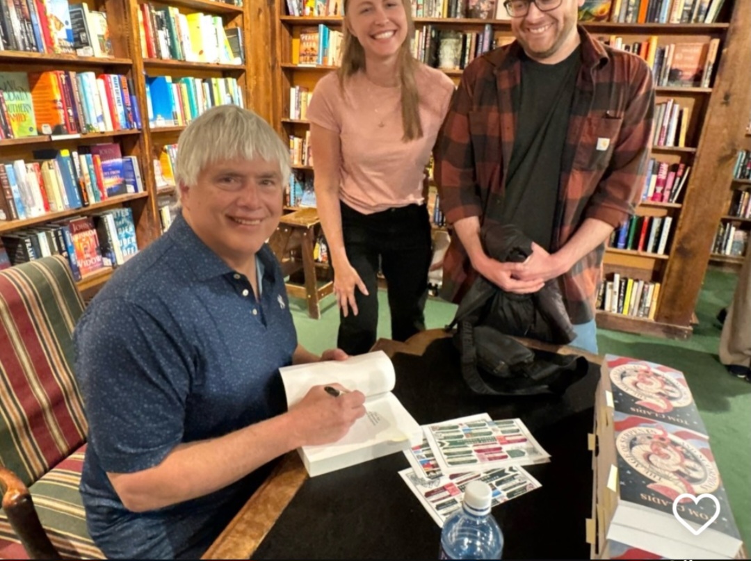 Three people in a bookstore, one seated signing a book, two standing smiling, bookshelves in background.