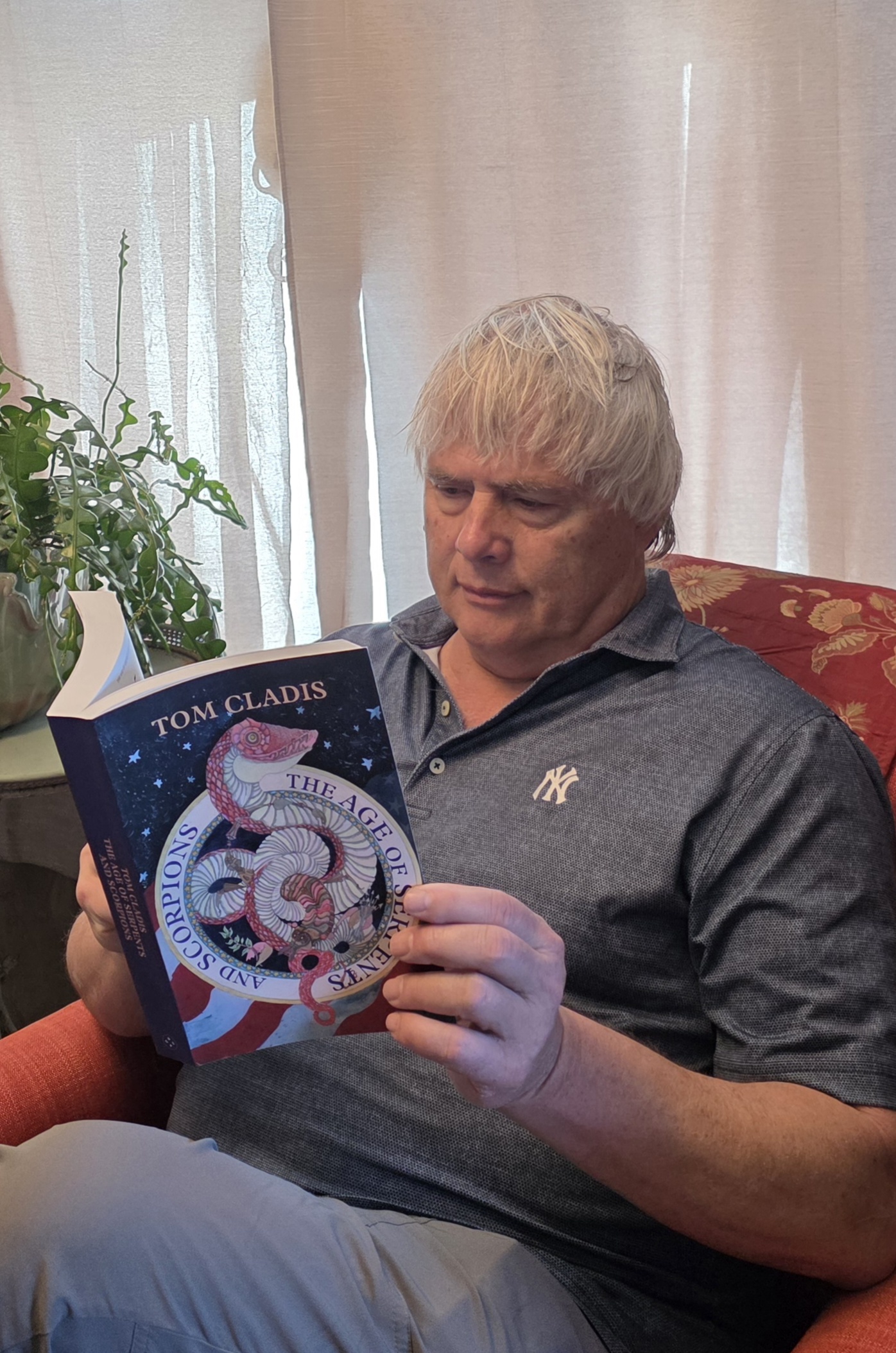 Older man with gray hair reading a book titled 'Tom Cladis' in a room with curtains and a potted plant.