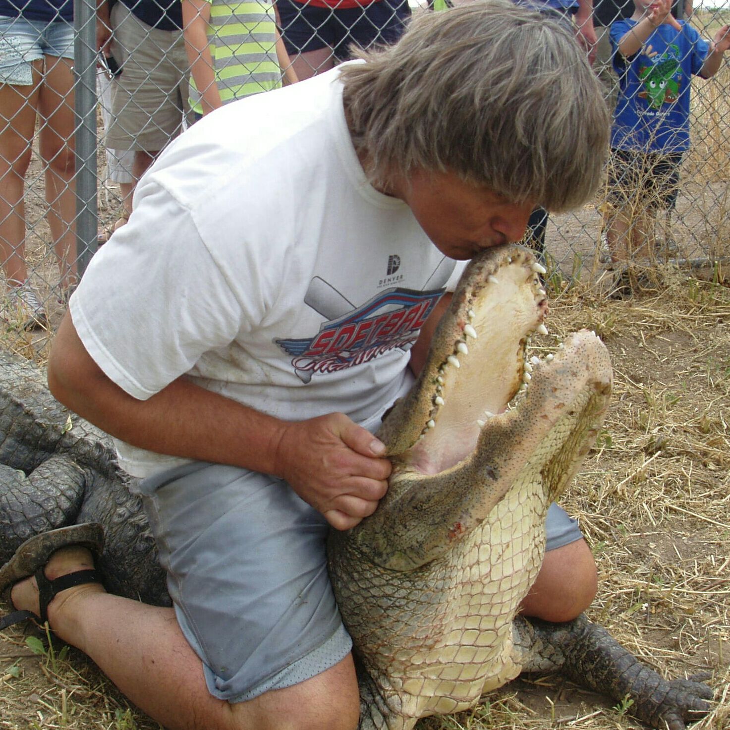 Man kneeling and kissing the open mouth of a crocodile outdoors, with a group of people behind a fence.